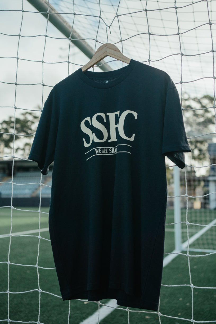 Black t-shirt with 'SSFC' and 'WE ARE SSFC' text on a hanger against a soccer goal background