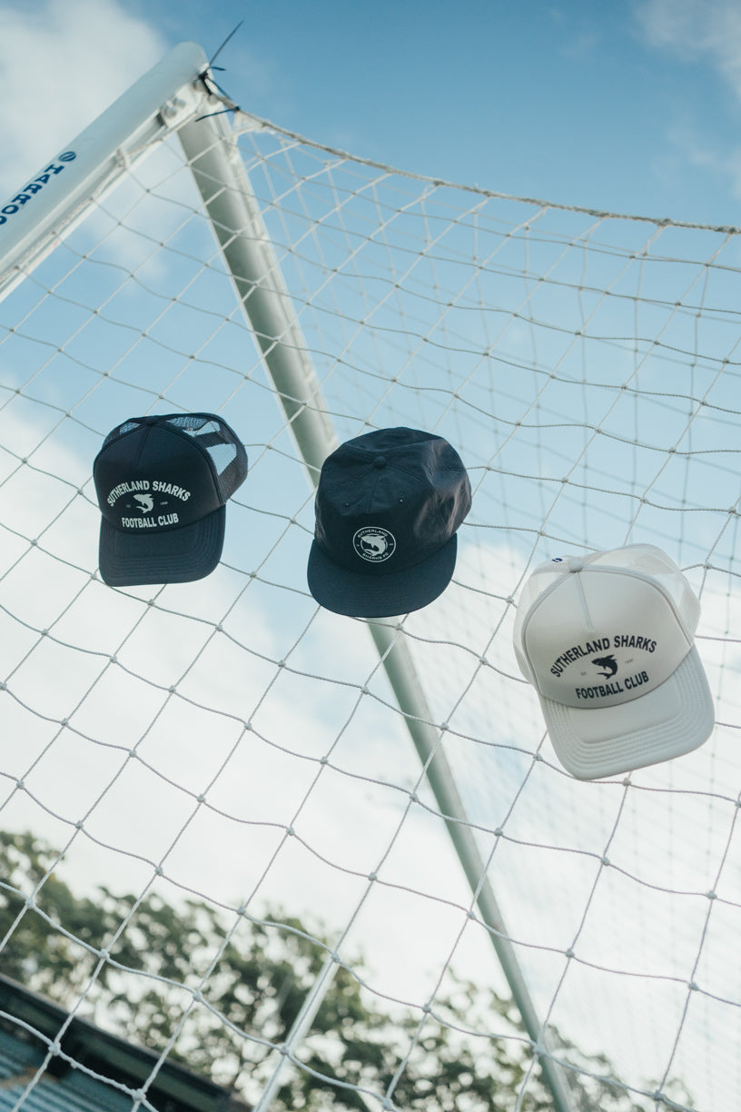 Three caps with a logo hanging on a soccer goal net against a blue sky.